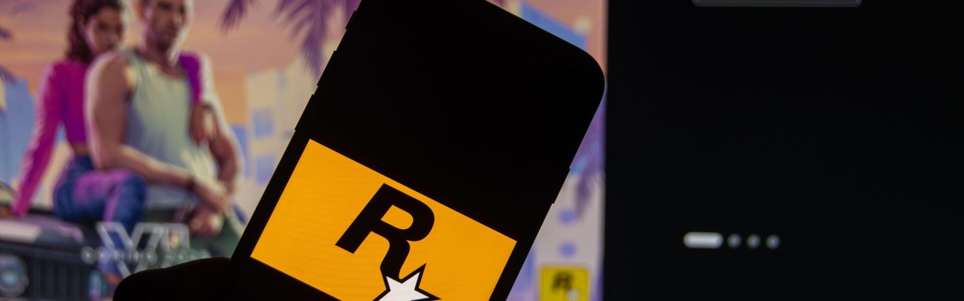 a person holding a phone with the Rockstar logo with GTA VI trailer in the background on a monitor
