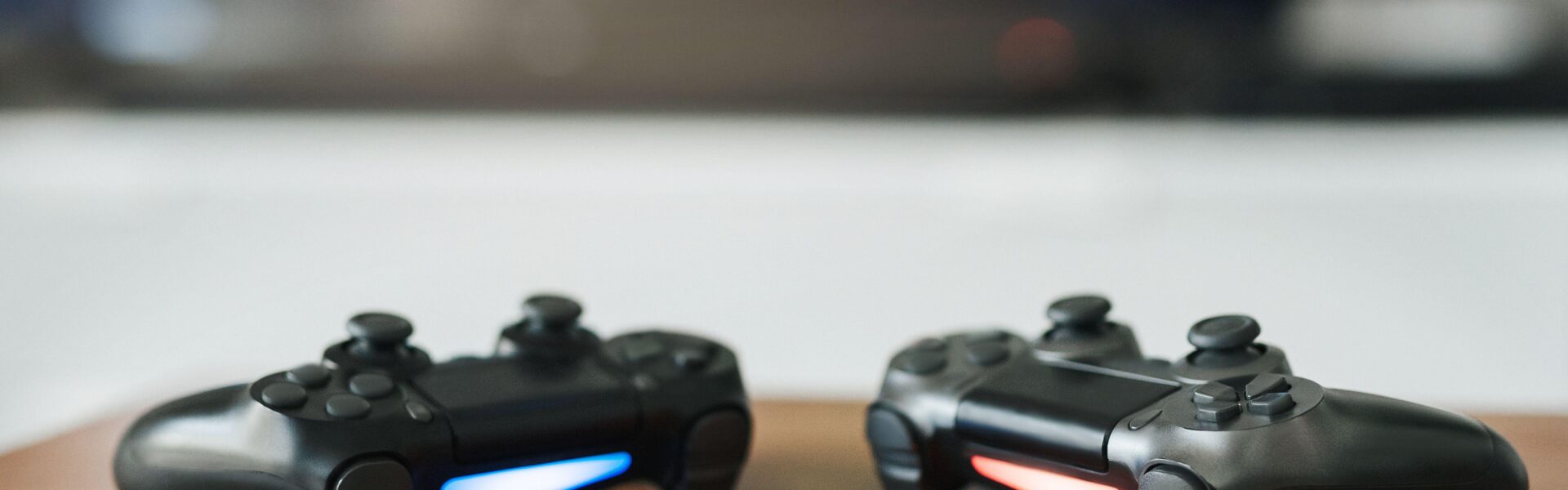 current console generation, Playstation 5 controllers on a wooden table with a game on the monitor behind them
