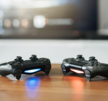 current console generation, Playstation 5 controllers on a wooden table with a game on the monitor behind them
