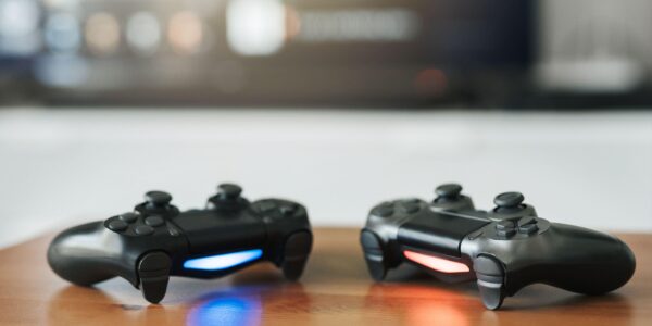 current console generation, Playstation 5 controllers on a wooden table with a game on the monitor behind them